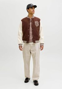 Brun varsityjakke med kremfargede ermer, med en logo, stripete krage og knappelukking. Brukt over en rutete skjorte og lyse bukser.