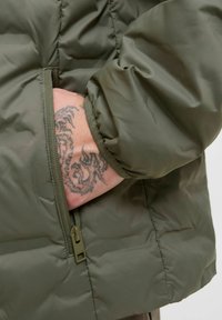 Ruka s tetováním draka částečně zasunutá do uzavřené kapsy rukávu olivově zelené prošívané bundy.