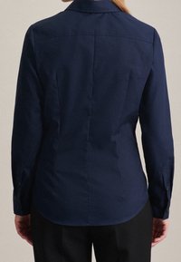 Kvinna som bär en åtsittande marinblå långärmad skjorta och svarta byxor, sedd bakifrån, stående mot en enkel ljus bakgrund.