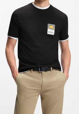 Homme portant un t-shirt noir avec un liseré blanc et un logo design soleil jaune et montagne au-dessus de "BOSS", associé à un pantalon beige et une ceinture noire.