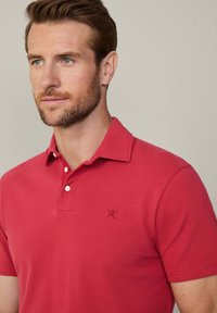 Červená polokošeľa s golierom, dvoma bielymi gombíkmi a malým vyšitým logom na ľavej hrudi. Mäkká textúra a priliehavý strih.