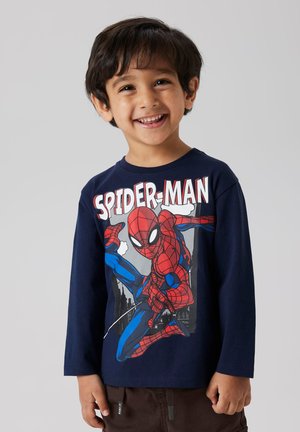 Un jeune garçon souriant portant une chemise à manches longues bleu marine avec un grand graphique coloré de Spider-Man et un pantalon marron.