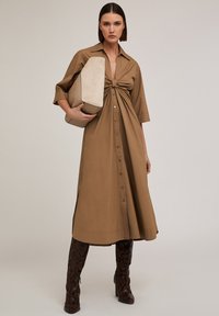 Abito camicia marrone con bottoni, nodo frontale, maniche corte e spacco laterale, abbinato a una borsa angolare beige e stivali con stampa serpente.