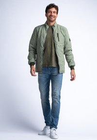 Helle grüne gesteppte Bomberjacke mit einem Reißverschlusstaschen auf der Brust und gerippten Bündchen, getragen über einem gedeckten olivfarbenen Hemd und blauen Jeans.