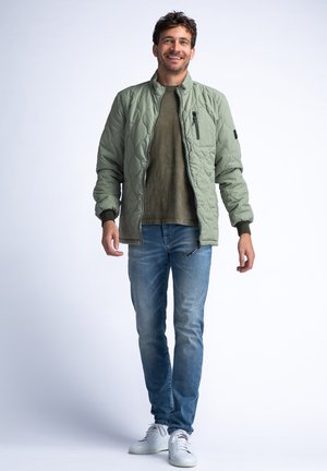 Helle grüne gesteppte Bomberjacke mit einem Reißverschlusstaschen auf der Brust und gerippten Bündchen, getragen über einem gedeckten olivfarbenen Hemd und blauen Jeans.