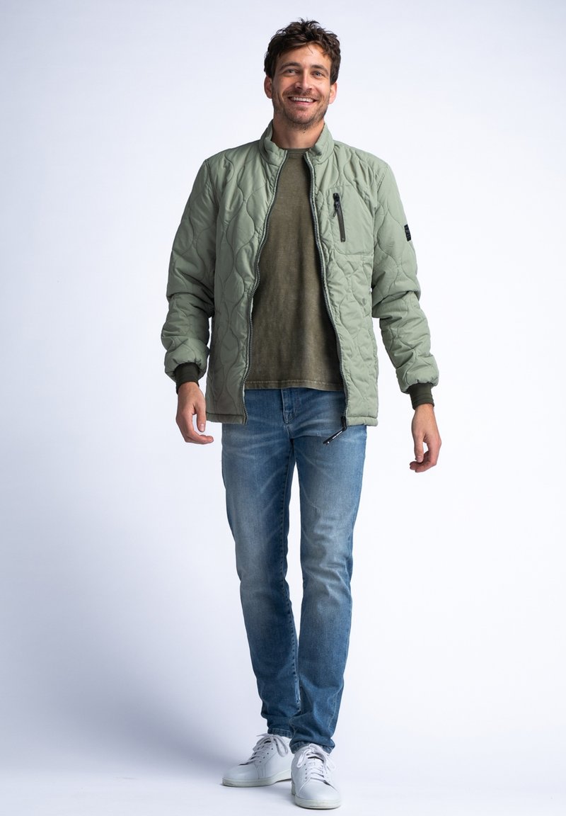 Helle grüne gesteppte Bomberjacke mit einem Reißverschlusstaschen auf der Brust und gerippten Bündchen, getragen über einem gedeckten olivfarbenen Hemd und blauen Jeans.