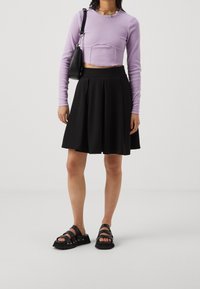 Crop top a maniche lunghe lilla, gonna plissettata nera e sandali neri con cinturini. Design semplici, texture morbide, con una vestibilità rilassata.