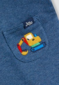 Sininen kangaspussi, jossa on kirjailtu keltainen ja punainen kaivinkone sekä pieni tummansininen etiketti, jossa lukee JoJo.