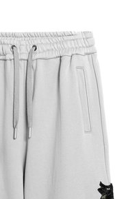 Pantalones de chándal gris claro con cintura elástica, cordón, bolsillos laterales y un diseño bordado en negro en la parte inferior de la pierna izquierda.