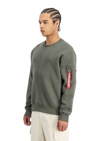 Sudadera de algodón verde con cuello redondo, mangas largas, puños acanalados y un bolsillo con cremallera que presenta una etiqueta roja en el brazo izquierdo.