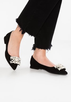 Zwarte platte schoenen met spitse neus en parel- en strassversieringen, gedragen met zwarte broek met rafelige zoom.