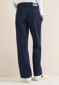 Jeans de mezclilla oscura de pierna ancha con dos bolsillos traseros, franjas laterales blancas y dobladillo con costura, combinados con zapatillas blancas.