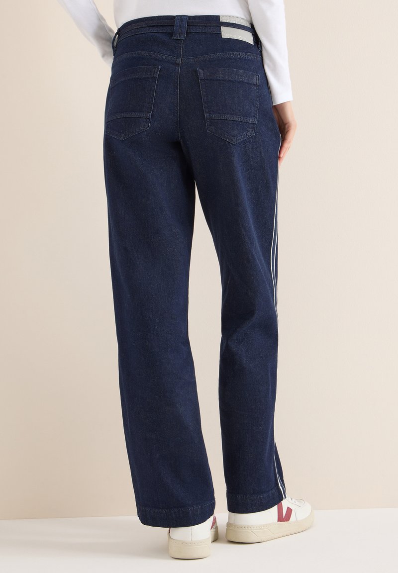 Jeans de mezclilla oscura de pierna ancha con dos bolsillos traseros, franjas laterales blancas y dobladillo con costura, combinados con zapatillas blancas.