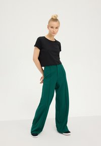 Maglietta nera corta abbinata a pantaloni larghi a vita alta verdi. I pantaloni presentano pince frontali e una texture liscia. Sneakers bianche completano il look.