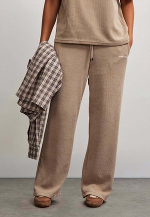 Persona che indossa pantaloni beige larghi a coste e scarpe marroni, tenendo in una mano una camicia beige e nera a quadri piegata.