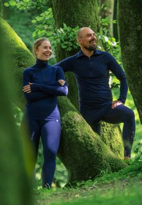 Två personer i åtsittande marinblå friluftskläder, en i en zip-up tröja, står mot mossklädda träd i en frodig grön miljö.