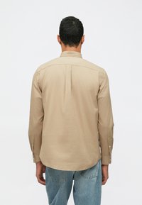 Uomo in piedi di spalle, indossa una camicia beige a maniche lunghe e jeans azzurri chiari, su uno sfondo bianco semplice.