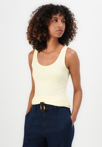 Mujer con cabello rizado, vestida con una blusa sin mangas de un amarillo pálido y pantalones de cintura alta en azul oscuro con una hebilla de cinturón de carey.