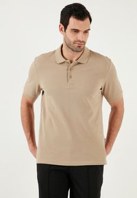 Béžové polo tričko s krátkým rukávem, s knoflíkovým zapínáním na tři knoflíky a texturovanou látkou. V kombinaci s černými kalhotami, s jednoduchým designem bez vzorů.
