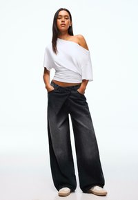 Top bianco oversize con scollo a barca abbinato a jeans neri a gamba larga; tessuto texturizzato con finitura sbiadita, dotato di tasche laterali e cintura elastica.