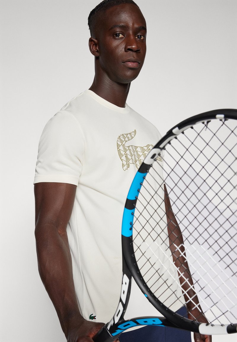 T-shirt créme à manches courtes avec un design de logo doré texturé, associé à une raquette de tennis noire et bleue, sur un fond neutre.