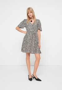 Ξανθιά γυναίκα στέκεται με το χέρι στη μέση, φορώντας ένα κοντομάνικο μαύρο και λευκό πουά φόρεμα και μαύρες χαμηλοτάκουνες εσπαντρίγιες.