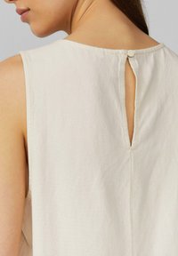 Mouwloze top van lichtbeige stof met een sleutelgat aan de achterkant, voorzien van een enkele knoopsluiting en zichtbare stikseldetails.