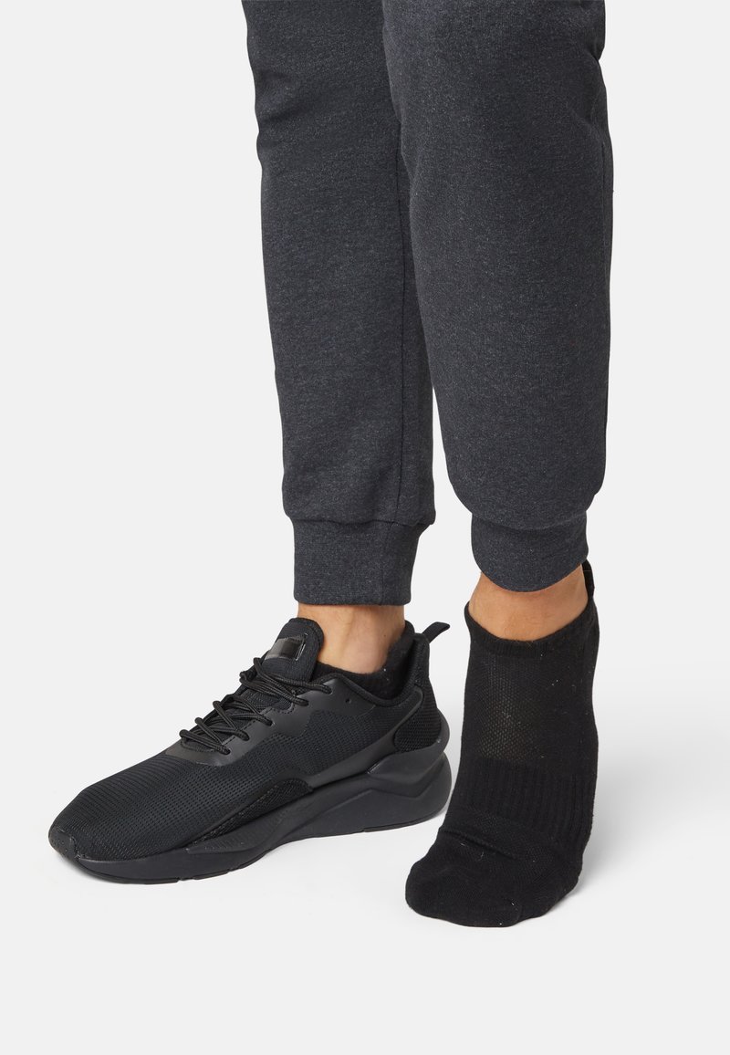 Black athletic sneaker with mesh upper and rubber sole, alongside a black sock with ribbed and sheer sections on a gray jogger.