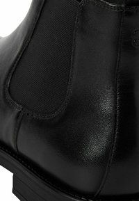 Botín Chelsea de cuero negro con paneles laterales elásticos, diseño elegante, textura suave y tacón de goma bajo. Se aprecian detalles de costura.