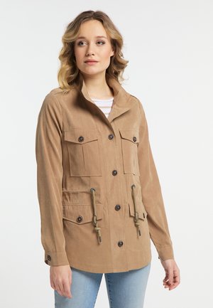 Vrouw met schouderlang blond haar, draagt een beige jasje met knopen en vier voorzakken, en lichtblauwe spijkerbroek, kijkt iets naar rechts.