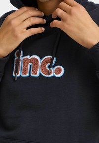 Sudadera navy con cordones, que presenta el texto bordado "inc." en marrón y azul claro. Tejido suave y corte relajado.