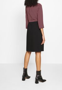 Robe noire avec un haut maroon à motifs, manches trois-quarts et silhouette ajustée. Portée avec des bottines noires brillantes.