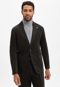 Dunkelbrauner Cordblazer mit schmalem Revers, zwei Knöpfen und Fronttaschen. Über einem grauen Rollkragenpullover getragen. Glatte Textur.