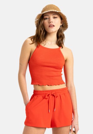 Rode geribde crop top met golvende zoom, gecombineerd met bijpassende rode short met trekkoord. Het model draagt ook een rieten bucket hat.