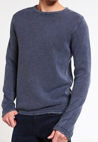 Mann, der einen anliegenden blauen strukturierten Strickpullover mit langen Ärmeln und gerippten Bündchen trägt, kombiniert mit dunklen Hosen.