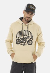 Un hoodie beige avec un graphique de moto noir et le texte "VON DUTCH". Il a une poche avant et une capuche avec cordon de serrage. Porté avec un jean foncé et une casquette.