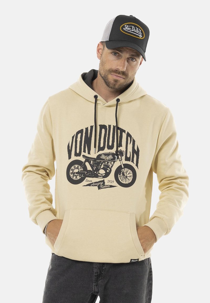 Un hoodie beige avec un graphique de moto noir et le texte "VON DUTCH". Il a une poche avant et une capuche avec cordon de serrage. Porté avec un jean foncé et une casquette.