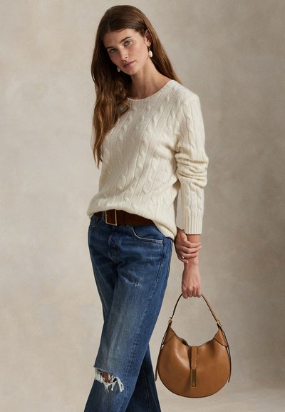 Frau in cremefarbenem Zopfmuster-Pullover und zerrissenen blauen Jeans hält eine runde braune Ledertasche vor neutralem Hintergrund.