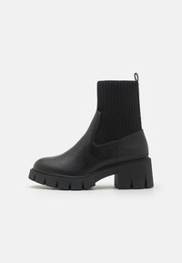 Black ankle boot with a ribbed knit upper, smooth leather body, block heel, and chunky rubber outsole. Simple, modern design.