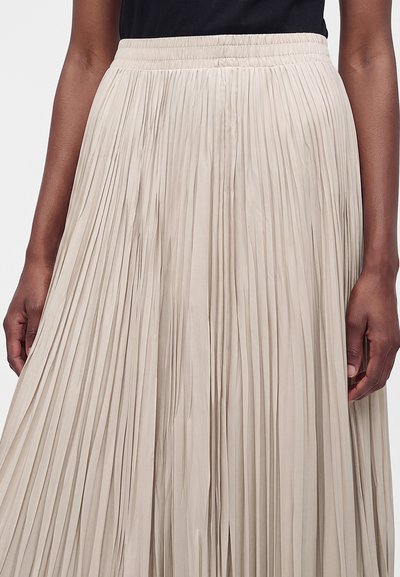 Femme portant une jupe plissée beige avec une taille élastique et un haut noir sans manches, les mains reposant sur les côtés.