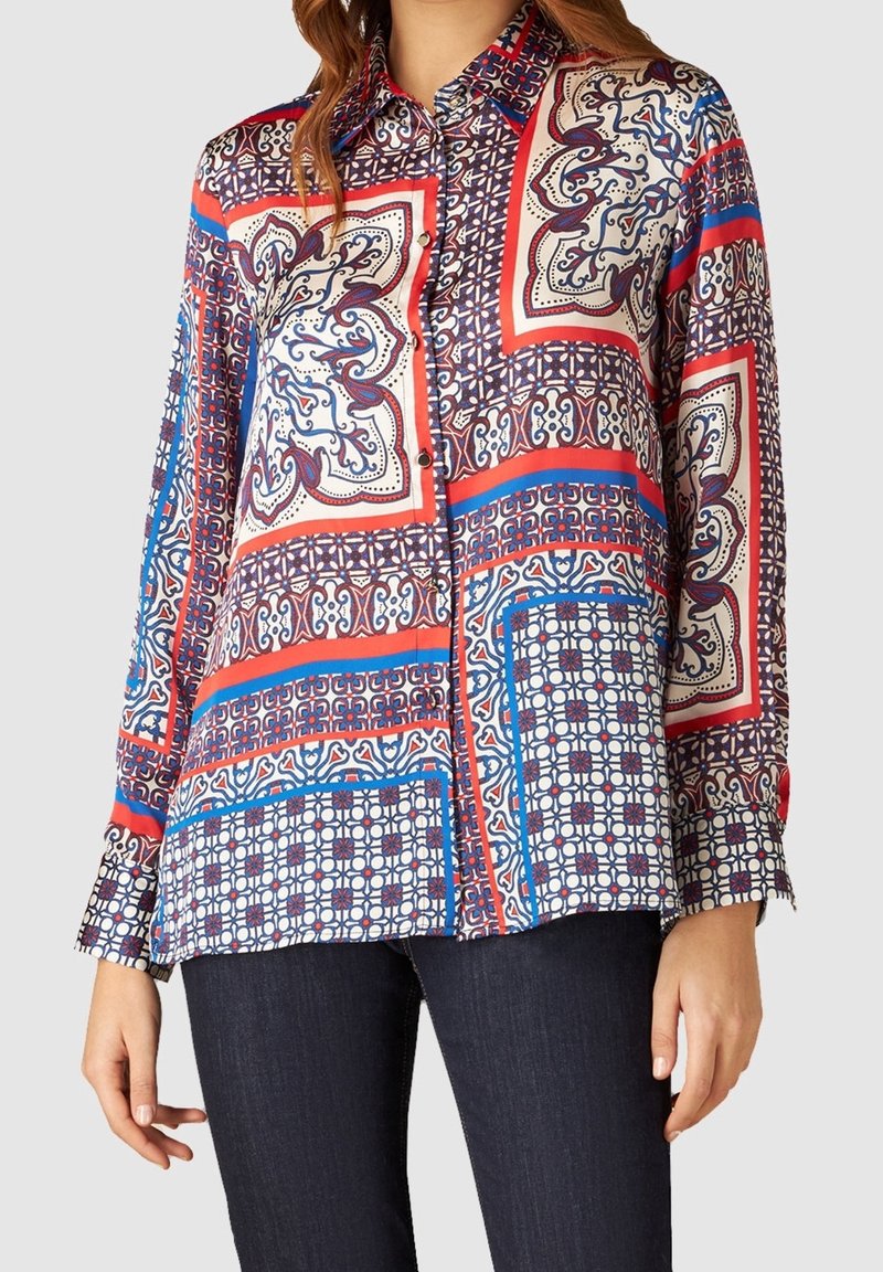 Blusa a maniche lunghe con una stampa geometrica multicolore in blu, rosso e crema. Tessuto leggero e morbido; chiusura con bottoni sul davanti e colletto.