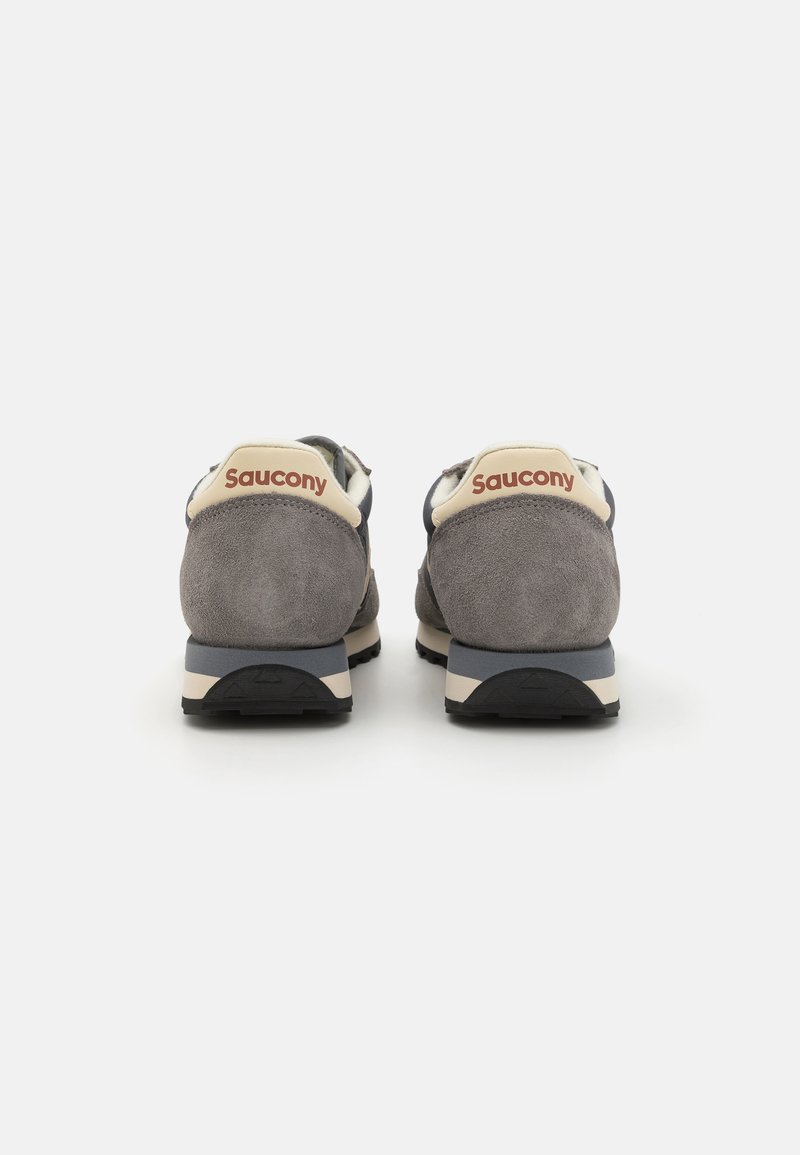 Le sneakers in suede grigio presentano un tallone in crema con il marchio "Saucony", accenti blu scuro e una suola nera texturizzata.