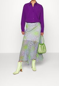 Lila Hemd, durchsichtiger gemusterter Rock in Grün und Lavendel, grüne Netzhandtasche und hellgrüne Absatzstiefel mit seitlichen Reißverschlüssen.