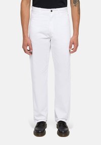 Hombre vestido con pantalones rectos blancos y zapatos negro con cordones, de pie contra un fondo claro y liso.