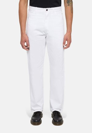 Homme portant un pantalon blanc à jambes droites avec des chaussures noires à lacets, se tenant devant un fond clair uni.