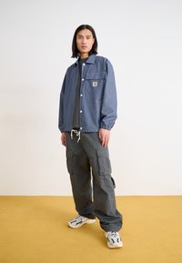 Denim jacket with snap buttons and chest pocket, paired with olive cargo pants featuring multiple pockets, and chunky sneakers.