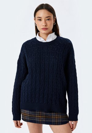 Navyblauw kabelgebreid sweatshirt met een ronde halslijn en ribgebreide mouwen, gecombineerd met een geruite rok met gele en witte accenten.