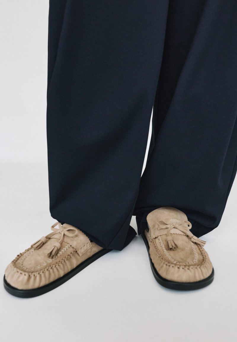 Chaussons mocassins en daim beige à pompons, portés avec un pantalon ample et foncé, pieds positionnés proches l’un de l’autre sur fond blanc.