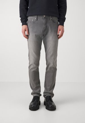 Hombre con vaqueros gris desgastados de corte ajustado, zapatos negros con cordones y una camiseta negra de manga larga, de pie sobre un fondo gris claro.