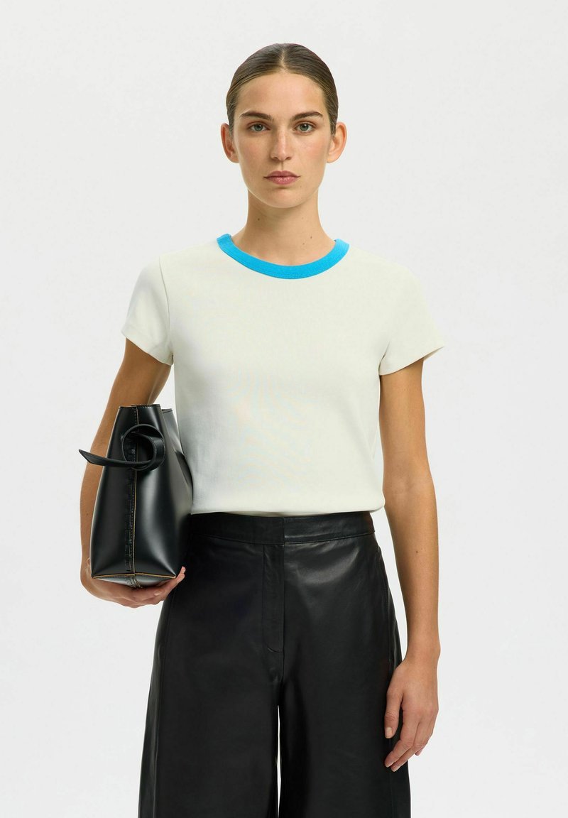 Haut blanc à manches courtes avec un col bleu, associé à un pantalon en cuir noir taille haute. Le mannequin tient un sac noir avec une poignée circulaire.